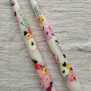 Könnte beinhalten: Zwei weiße Spitzkerzen mit handgemalten Blumenmustern. Die Blumen sind in Rosa-, Gelb-, Rot- und Blautönen gehalten, mit grünen Blättern. Die Kerzen stehen auf einem neutralfarbenen Stoff.