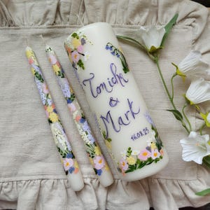 May include: Three white candles, two tapers and a pillar candle, adorned with hand-painted floral patterns in pastel hues. The pillar candle is inscribed with the names "Tonicha & Mark" and the date "16.08.25".