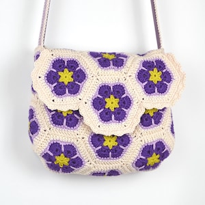 May include: A handmade crochet crossbody bag featuring a floral pattern. The bag is cream-colored with purple and yellow flower motifs. It has a scalloped flap and a matching strap, perfect for carrying essentials.
