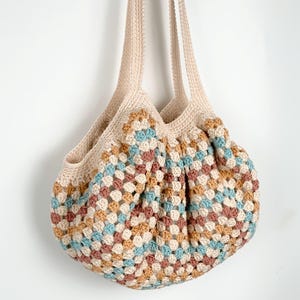May include: A handmade crochet tote bag in a granny square pattern. The bag features a cream-colored top and straps, with a body of colorful squares in shades of blue, brown, tan, and cream. The bag is suspended against a white background.