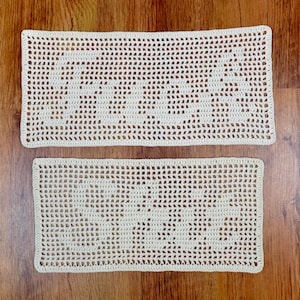 Peut inclure: Deux napperons rectangulaires en crochet blanc cassé sont présentés sur une surface en bois. Les napperons présentent un motif ajouré et répétitif. Le plus grand est au-dessus du plus petit, tous deux mettant en valeur une esthétique artisanale.