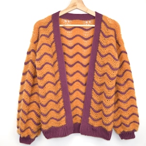 Peut inclure: Un cardigan en crochet à manches longues avec un motif ondulé jaune moutarde et violet. Le vêtement présente une bordure côtelée violette unie le long de l'ouverture avant, des poignets et de l'ourlet, avec une texture en maille ouverte.