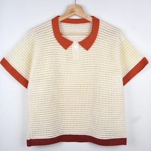 Peut inclure: Un chemisier à manches courtes en crochet blanc avec un col et des bordures orange. Le chemisier a un col boutonné et une coupe ample.