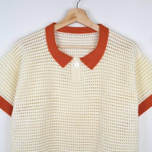 Pode incluir: Polo creme de malha aberta, com mangas curtas e gola e acabamentos das mangas laranja. A camisa tem uma carcela abotoada e est&aacute; pendurada num cabide de madeira. A textura do croch&ecirc; &eacute; vis&iacute;vel.