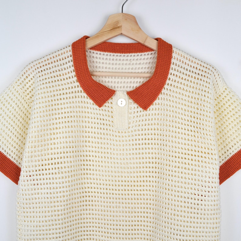 Pode incluir: Polo creme de malha aberta, com mangas curtas e gola e acabamentos das mangas laranja. A camisa tem uma carcela abotoada e est&aacute; pendurada num cabide de madeira. A textura do croch&ecirc; &eacute; vis&iacute;vel.