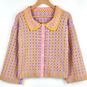 Puede incluir: Un cárdigan de ganchillo con cierre de botones y un cuello de contraste rosa y amarillo. El cárdigan está hecho con hilo de color morado claro y amarillo.