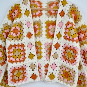 May include: A white crocheted cardigan with a granny square pattern in shades of pink, orange, and brown.