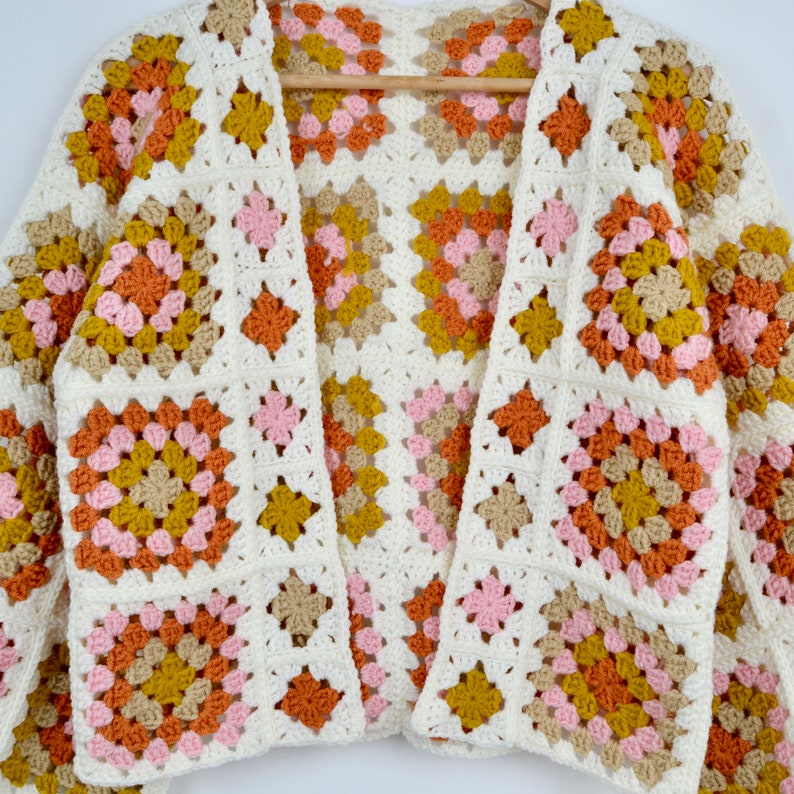 May include: A white crocheted cardigan with a granny square pattern in shades of pink, orange, and brown.