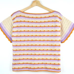 Peut inclure: Un haut en crochet à manches courtes avec des rayures horizontales crème, violet clair et orange vif. Le vêtement présente une large encolure bateau et un motif de points texturé sur l'ensemble, avec des manches crème et une bordure orange.