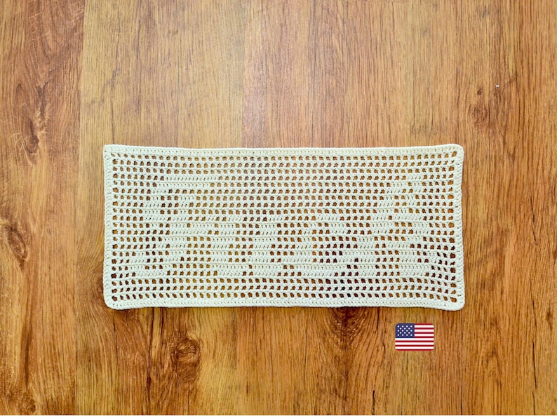 Puede incluir: Tapete rectangular de ganchillo color crema. El dise&ntilde;o calado presenta un patr&oacute;n repetitivo de peque&ntilde;os agujeros espaciados uniformemente. El tapete est&aacute; colocado sobre una superficie de madera, con la bandera americana en la esquina inferior derecha.
