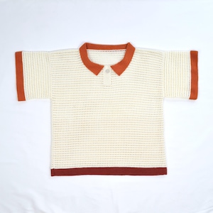 Pode incluir: Camisa polo creme, de manga curta, com um design de malha texturizada. A gola, os punhos e a bainha apresentam uma borda cor de ferrugem. A camisa tem uma carcela abotoada no decote.