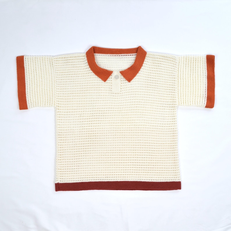 Pode incluir: Camisa polo creme, de manga curta, com um design de malha texturizada. A gola, os punhos e a bainha apresentam uma borda cor de ferrugem. A camisa tem uma carcela abotoada no decote.