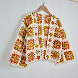May include: A white crocheted cardigan with a granny square pattern in shades of pink, orange, and brown. The cardigan has a long, open front and long sleeves.