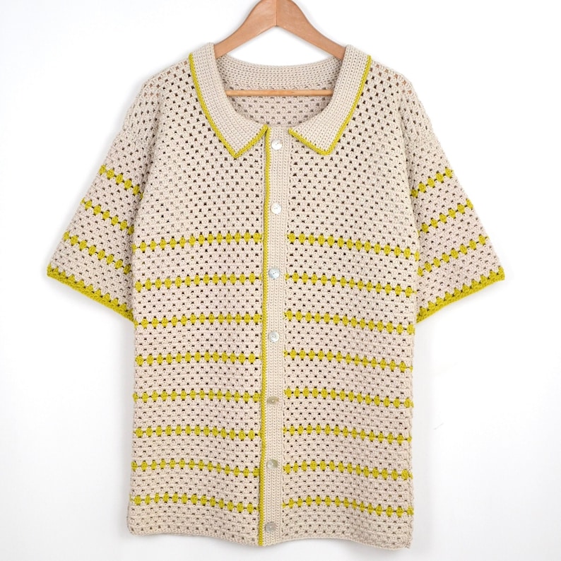 Puede incluir: Una camisa de crochet color crema, de manga corta, con cuello y cierre de botones en la parte delantera. La camisa presenta rayas horizontales y ribetes en un color amarillo-verdoso brillante. La camisa est&aacute; colgada en una percha de madera.