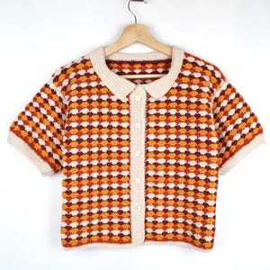 Può includere: Una camicia all'uncinetto a maniche corte con bottoni in un motivo ripetuto di arancione, crema e bordeaux. La camicia ha un colletto e finiture color crema ed è appesa a una gruccia di legno. Il design all'uncinetto crea un aspetto strutturato e fantasia.