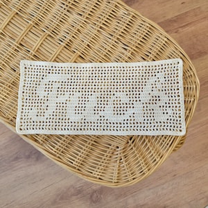 Puede incluir: Tapete rectangular de ganchillo color crema con un dise&ntilde;o calado. El tapete est&aacute; colocado sobre una superficie de mimbre tejido, con un suelo de madera visible en el fondo. El patr&oacute;n de ganchillo presenta una serie de peque&ntilde;os agujeros espaciados.