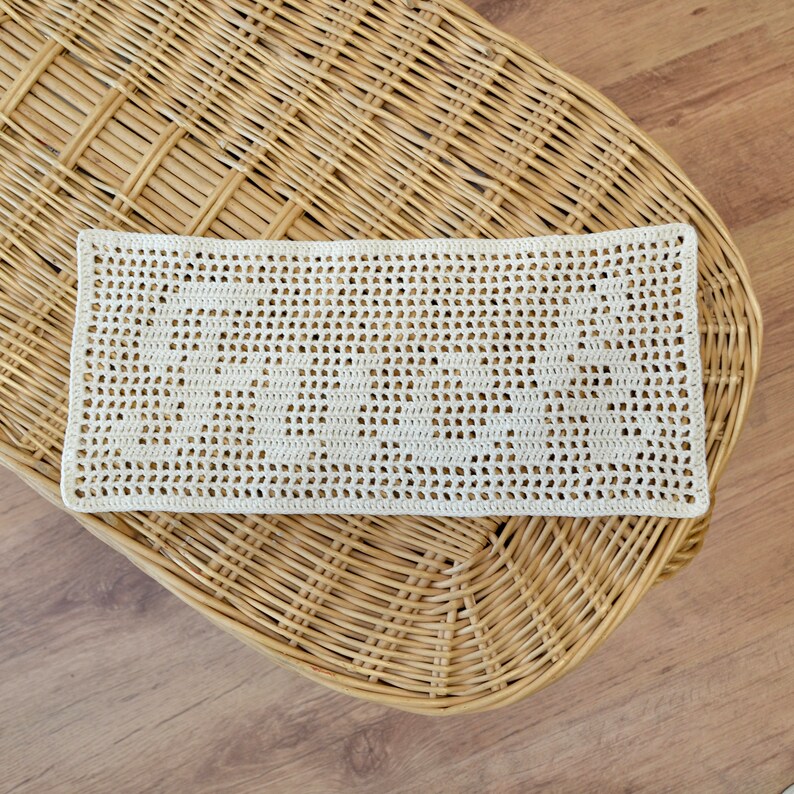 Puede incluir: Tapete rectangular de ganchillo color crema con un dise&ntilde;o calado. El tapete est&aacute; colocado sobre una superficie de mimbre tejido, con un suelo de madera visible en el fondo. El patr&oacute;n de ganchillo presenta una serie de peque&ntilde;os agujeros espaciados.