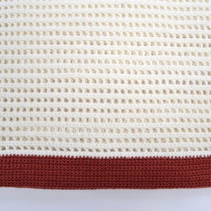 Pode incluir: Close-up de um cobertor de croch&ecirc; creme com um design de tecido aberto e texturizado. A borda inferior apresenta uma faixa s&oacute;lida de croch&ecirc; marrom avermelhado. O padr&atilde;o do cobertor cria uma apar&ecirc;ncia de grade, adequada para decora&ccedil;&atilde;o de casa.