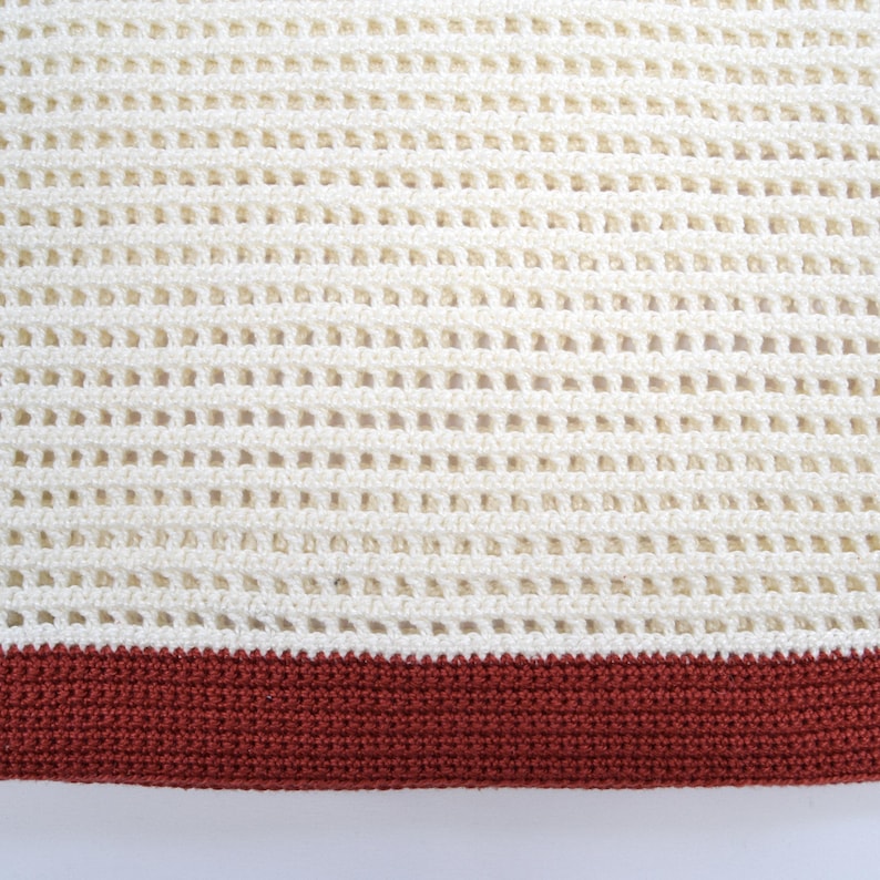 Pode incluir: Close-up de um cobertor de croch&ecirc; creme com um design de tecido aberto e texturizado. A borda inferior apresenta uma faixa s&oacute;lida de croch&ecirc; marrom avermelhado. O padr&atilde;o do cobertor cria uma apar&ecirc;ncia de grade, adequada para decora&ccedil;&atilde;o de casa.