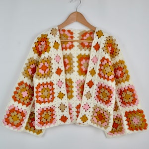 May include: A white crochet cardigan with a granny square pattern in shades of pink, orange, and brown. The cardigan has a V-neck and long sleeves.
