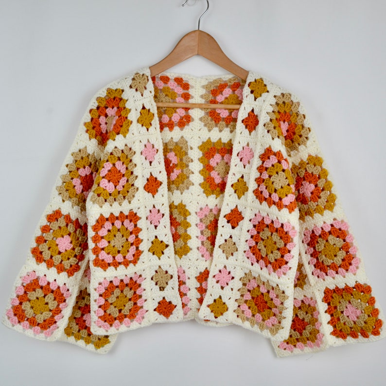 May include: A white crochet cardigan with a granny square pattern in shades of pink, orange, and brown. The cardigan has a V-neck and long sleeves.
