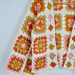 May include: A white crocheted cardigan with a granny square pattern in shades of pink, orange, and brown.