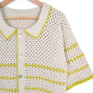 Puede incluir: Una camisa de crochet color crema con tejido abierto, cuello y mangas cortas. La camisa presenta un ribete amarillo-verdoso en el cuello y las mangas, con un patr&oacute;n horizontal del mismo color. Tiene tres botones transparentes en la parte delantera y est&aacute; colgada de una percha de madera.
