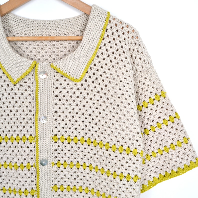 Puede incluir: Una camisa de crochet color crema con tejido abierto, cuello y mangas cortas. La camisa presenta un ribete amarillo-verdoso en el cuello y las mangas, con un patr&oacute;n horizontal del mismo color. Tiene tres botones transparentes en la parte delantera y est&aacute; colgada de una percha de madera.