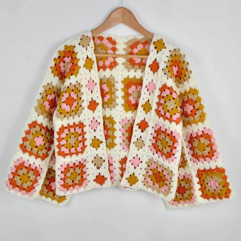 May include: A white crocheted cardigan with a geometric pattern in shades of pink, orange, and brown. The cardigan has a V-neck and long sleeves.