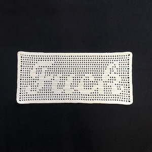 Puede incluir: Un tapete rectangular de ganchillo blanco roto con un delicado dise&ntilde;o calado. La pieza hecha a mano se muestra sobre un fondo negro, resaltando la intrincada textura y el patr&oacute;n del hilo. Los bordes del tapete est&aacute;n cuidadosamente acabados.