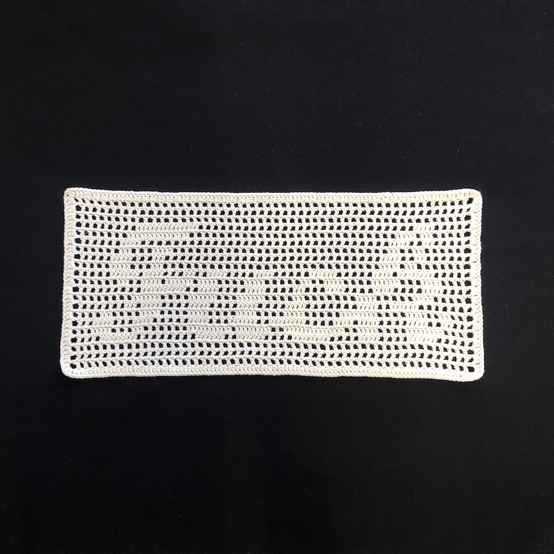 Puede incluir: Un tapete rectangular de ganchillo blanco roto con un delicado dise&ntilde;o calado. La pieza hecha a mano se muestra sobre un fondo negro, resaltando la intrincada textura y el patr&oacute;n del hilo. Los bordes del tapete est&aacute;n cuidadosamente acabados.