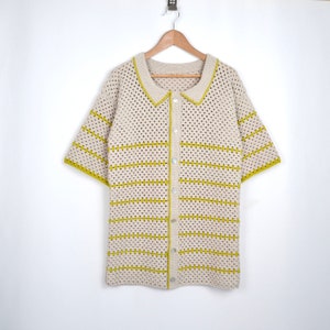Puede incluir: Una camisa de crochet color crema de manga corta con cuello. La camisa presenta rayas y ribetes horizontales amarillos. Se abrocha por delante con botones plateados y cuelga de una percha de madera.