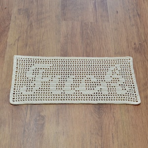 Puede incluir: Un tapete rectangular de ganchillo blanco roto con un patr&oacute;n calado repetitivo. La pieza hecha a mano se exhibe sobre una superficie de madera, mostrando su delicada textura y dise&ntilde;o. Los bordes del tapete est&aacute;n cuidadosamente acabados.