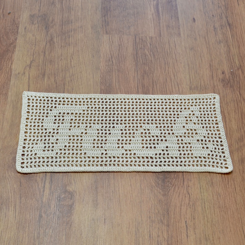 Puede incluir: Un tapete rectangular de ganchillo blanco roto con un patr&oacute;n calado repetitivo. La pieza hecha a mano se exhibe sobre una superficie de madera, mostrando su delicada textura y dise&ntilde;o. Los bordes del tapete est&aacute;n cuidadosamente acabados.
