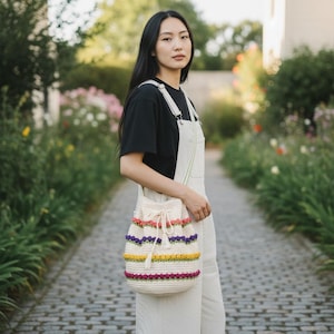 Peut inclure: Un sac seau en crochet de couleur crème orné de rangées horizontales de petits points bouffants en relief rappelant des fleurs, dans des tons rose, jaune et violet. Le sac possède une fermeture à cordon avec des pompons crème et une bandoulière.