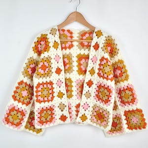 Peut inclure: Un cardigan en crochet crème à devant ouvert avec un motif de carrés granny. Les carrés présentent un motif de couleurs orange, rose et beige. Le cardigan est suspendu à une cintre en bois.