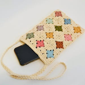 Pode incluir: Uma bolsa de telefone de crochê creme com um padrão floral colorido. A bolsa apresenta uma alça trançada e um telefone dentro. O padrão floral inclui quadrados rosa, azul, verde, laranja e marrom.