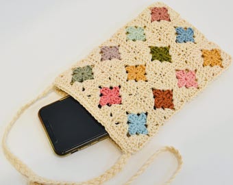 May include: A cream-colored crochet phone bag with a colorful floral pattern. The bag features a braided strap and a phone inside. The floral pattern includes pink, blue, green, orange, and brown squares.