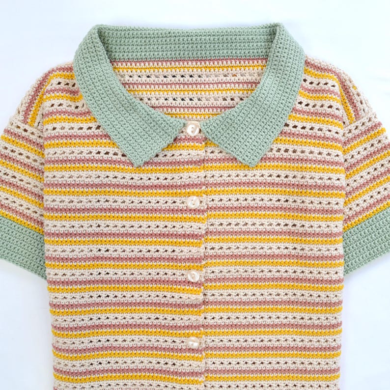 Puede incluir: Una camisa de crochet a rayas con un cuello verde y cierre de bot&oacute;n. La camisa tiene manga corta y est&aacute; hecha con hilo beige claro, amarillo y rosa.