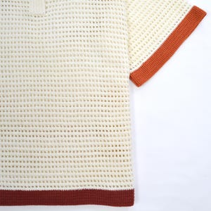 Pode incluir: Camiseta de croch&ecirc; creme com tecido aberto e mangas curtas. A camiseta apresenta uma gola polo e &eacute; adornada com uma borda marrom escura na bainha e punhos. O padr&atilde;o de croch&ecirc; cria um visual texturizado e respir&aacute;vel.