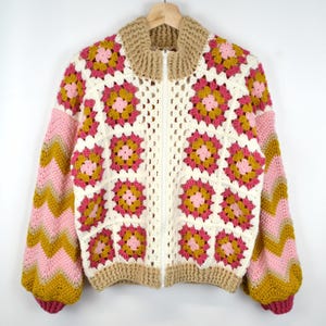 Peut inclure: Un cardigan en crochet blanc avec une fermeture éclair. Le cardigan a un col et des poignets marrons. Les manches sont à motif chevron rose et jaune.