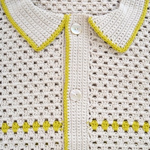 Puede incluir: Primer plano de un c&aacute;rdigan de crochet color crema con cuello y cierre de botones. Los bordes est&aacute;n ribeteados con hilo amarillo verdoso brillante. El c&aacute;rdigan presenta un patr&oacute;n decorativo en la parte inferior.