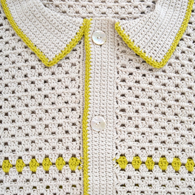 Puede incluir: Primer plano de un c&aacute;rdigan de crochet color crema con cuello y cierre de botones. Los bordes est&aacute;n ribeteados con hilo amarillo verdoso brillante. El c&aacute;rdigan presenta un patr&oacute;n decorativo en la parte inferior.