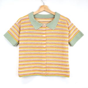 Puede incluir: Un top de crochet a rayas con cuello y mangas cortas. El top es en tonos de amarillo, rosa y blanco, con un ribete verde alrededor del cuello y las mangas.
