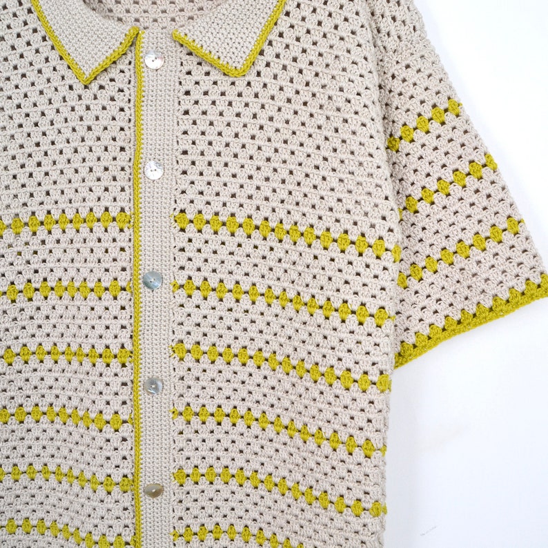 Puede incluir: Una camisa de crochet beige con mangas cortas y cuello. La camisa presenta rayas horizontales amarillas y se abrocha en la parte delantera con botones nacarados. Los bordes del cuello y las mangas est&aacute;n ribeteados en amarillo.