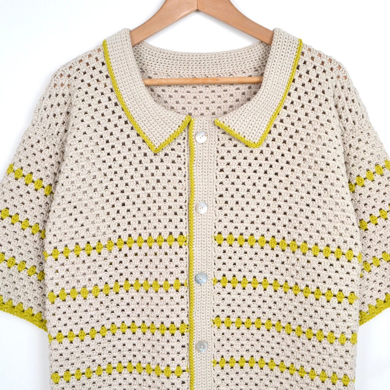 Puede incluir: Camisa de crochet color crema con cuello y mangas cortas. La camisa tiene una botonadura frontal y est&aacute; adornada con ribetes verde lima a lo largo del cuello, las mangas y las rayas horizontales. Cuelga de una percha de madera.
