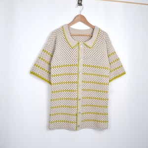 Puede incluir: Una camisa de crochet color crema de manga corta con botones en la parte delantera y cuello. La camisa presenta rayas horizontales y ribetes en un color verde amarillento brillante. La camisa est&aacute; colgada en una percha de madera sobre un fondo blanco.