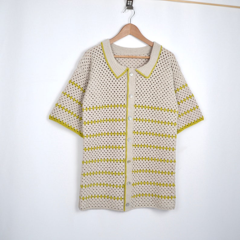 Puede incluir: Una camisa de crochet color crema de manga corta con botones en la parte delantera y cuello. La camisa presenta rayas horizontales y ribetes en un color verde amarillento brillante. La camisa est&aacute; colgada en una percha de madera sobre un fondo blanco.