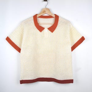 Pode incluir: Camisa polo creme de manga curta com design de croch&ecirc;. A camisa apresenta gola, acabamento das mangas e bainha laranja queimado. Um &uacute;nico bot&atilde;o branco fecha a gola. A camisa est&aacute; pendurada em um cabide de madeira sobre um fundo branco.