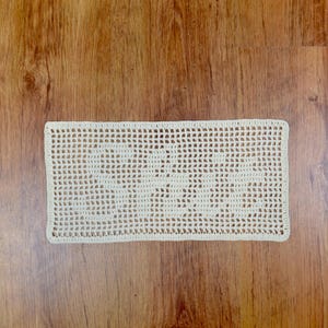 Puede incluir: Un tapete rectangular de ganchillo blanco roto con un delicado patr&oacute;n de tejido abierto. La pieza hecha a mano se muestra sobre una superficie de madera, mostrando su textura y dise&ntilde;o. Adecuado para la decoraci&oacute;n del hogar.